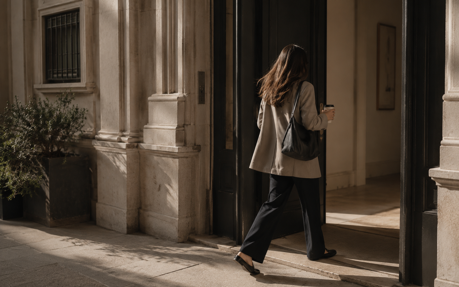 A younger professional seen from behind leaving home in understated modern workwear with a bag over her shoulder.