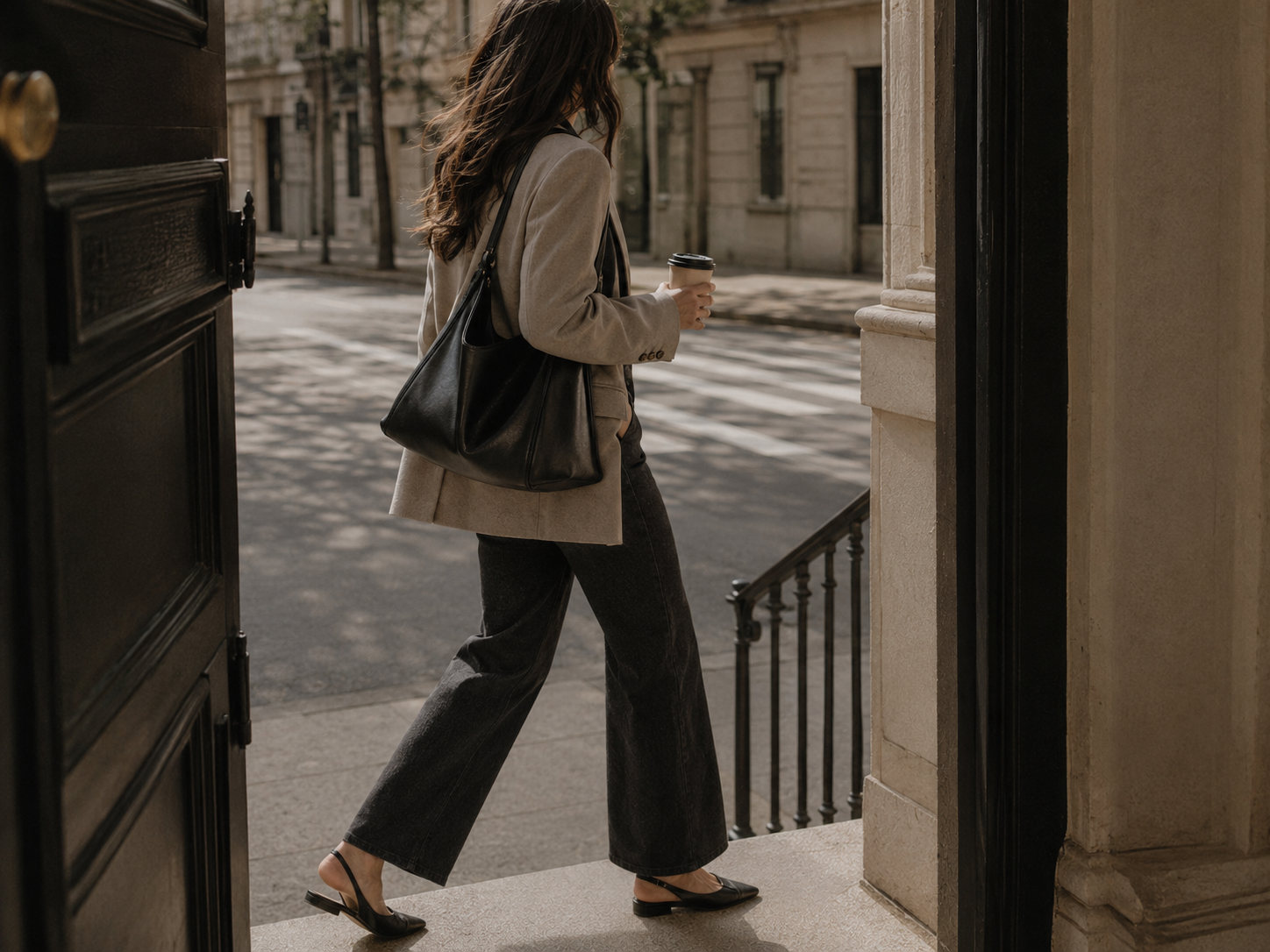 A younger professional in relaxed modern workwear walking through a city entrance with a shoulder bag.